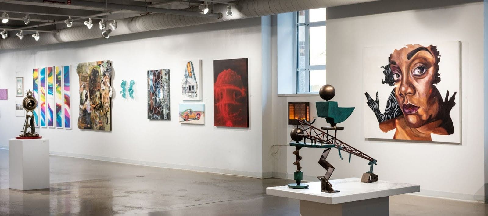 Gallery space featuring a metal sculpture in the foreground on the right corner, with two-dimensional artwork hanging on the wall behind it. Another sculpture is positioned in the background toward the far left center of the room.