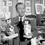 Black and white photo of Walt Disney inside a Disney studio filled with art, sketches, and puppets. He is holding up two wooden puppets and speaking directly to the camera.