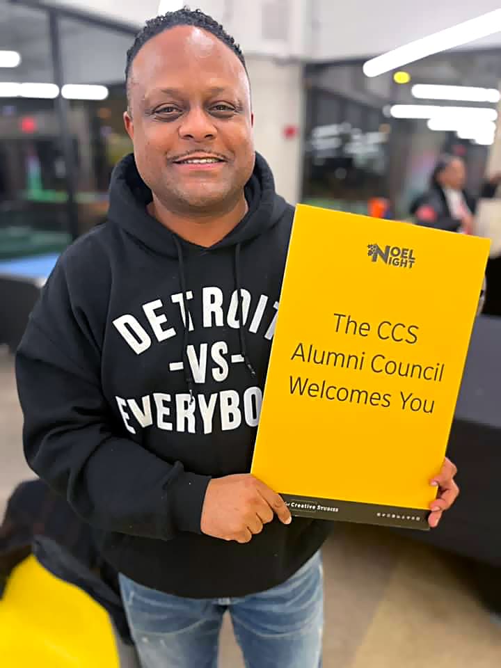 Alum in "Detroit vs Everybody" shirt holds a "The CCS Alumni council Welcomes You" sign.