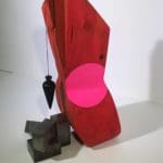 Abstract red sculpture with a black cone hanging off the side, above a few black blocks