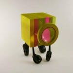 Abstract sculpture of a yellow and pink wooden block on wheels with a magnifying lens on one side